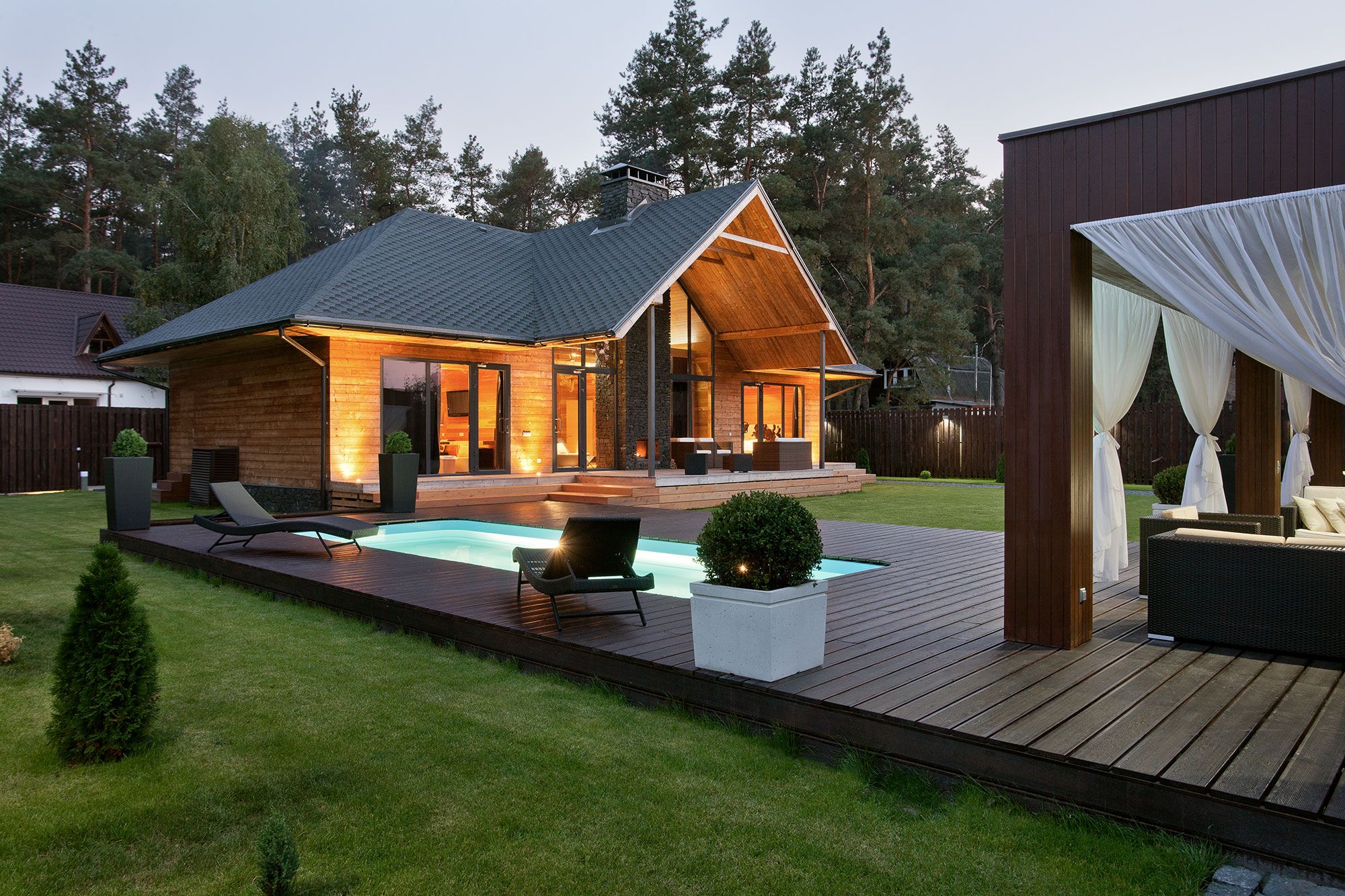 Modern timber home at twilight with glowing pool, wooden deck, lounge chairs, and pine trees behind