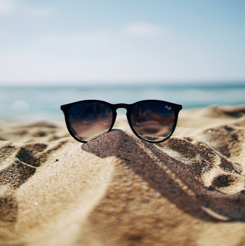 Pair of black round sunglasses resting on a sandy beach dune with blurred ocean and pale sky behind